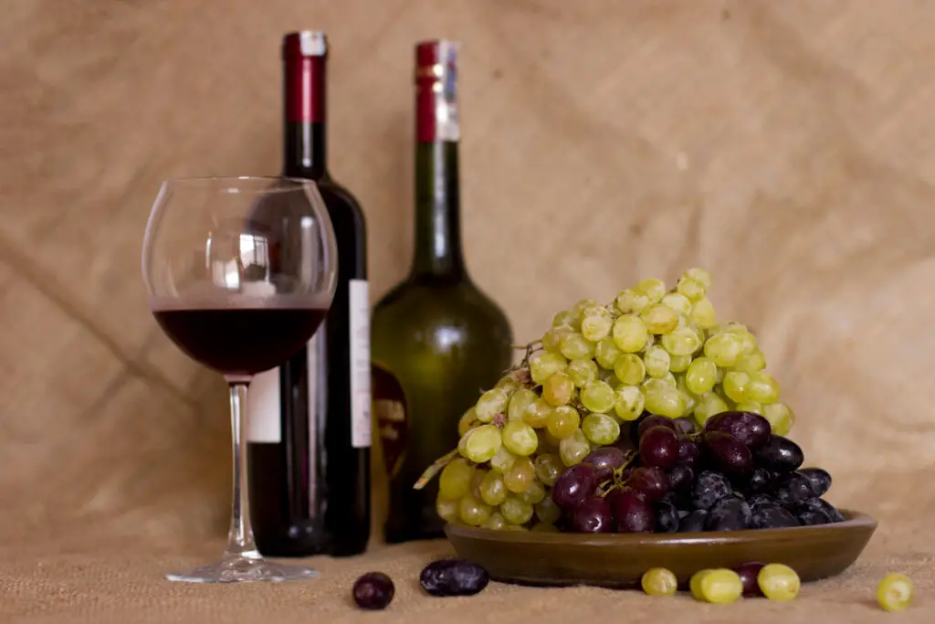 Blue and green grapes on the clay brown dish. Bottle with red and white wine on background