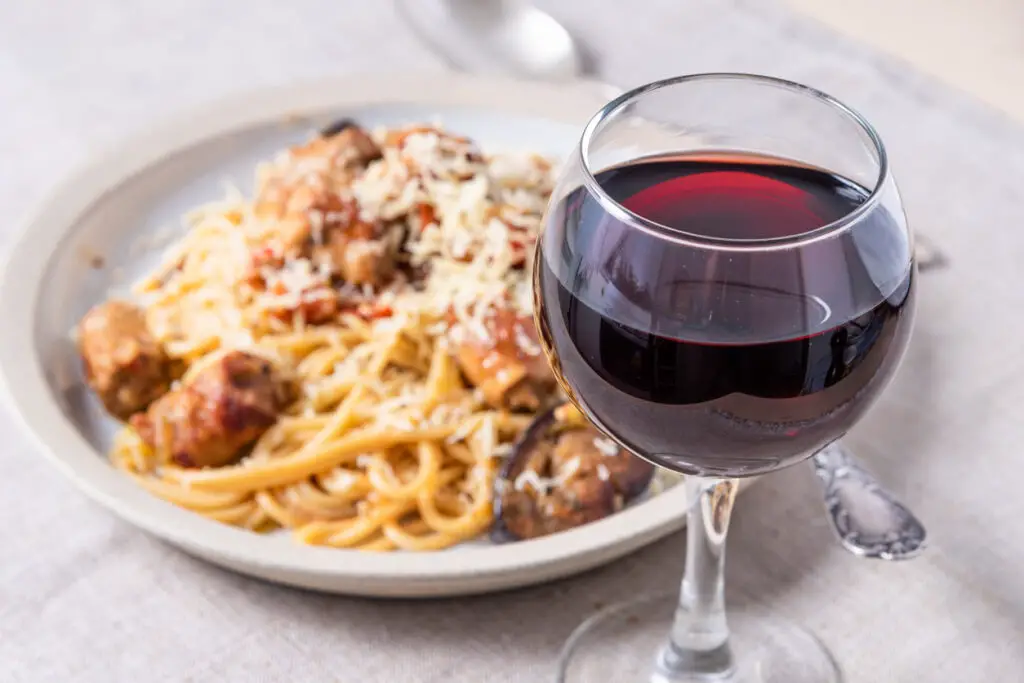 Spaghetti with homemade sausages and eggplant sprinkled with grated cheese in a plate and a glass of red wine in the foreground, selective focus - traditional Italian rural lunch
