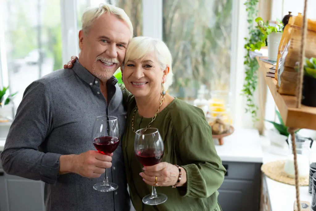 Cheerful silver-haired spouses cuddling and keeping glasses with red wine.