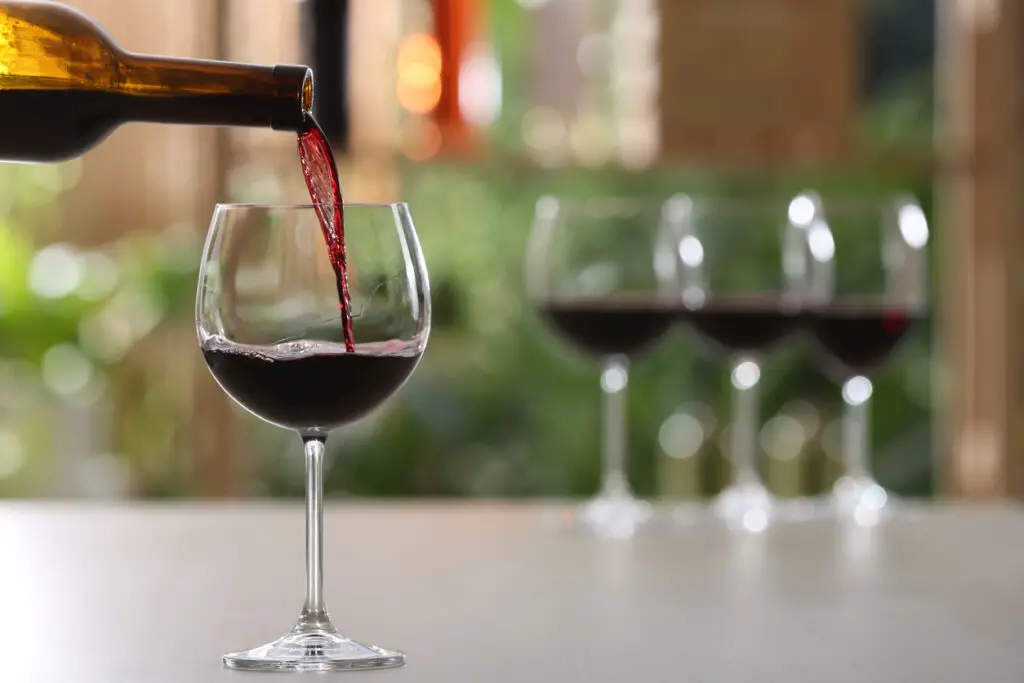 Pouring red wine from bottle into glass on table against blurred background, space for text