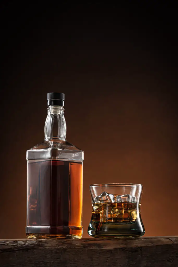 Bottle of whisky and glass of whisky with ice on wooden log with orange and black degraded background, with copy space.