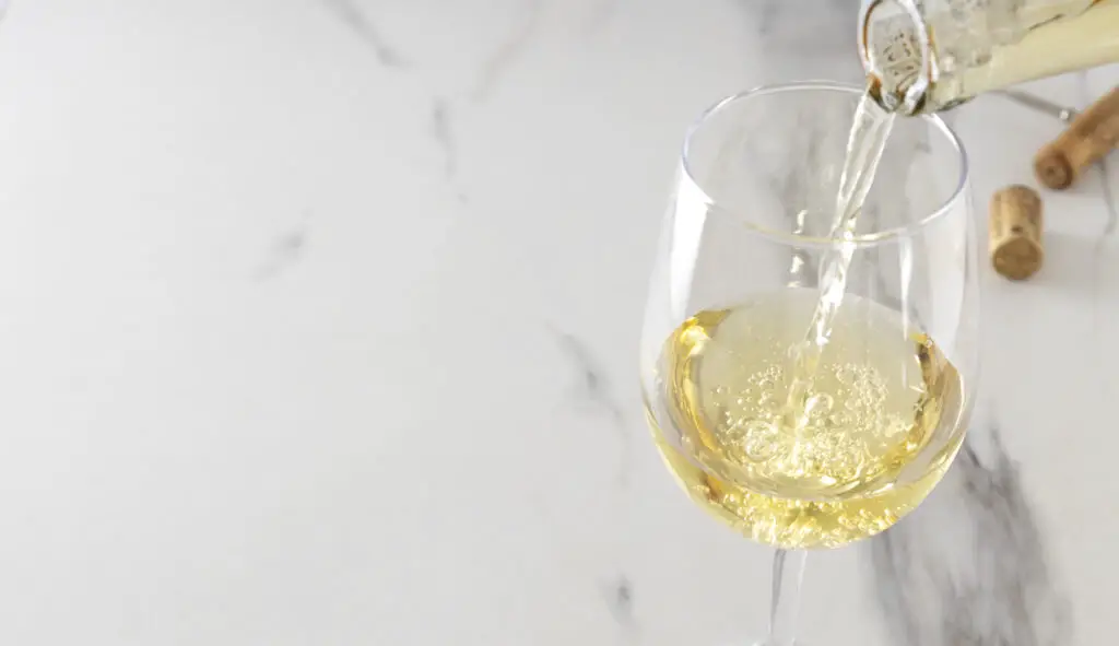 Closeup of of wine glass and process of pouring white wine, corkscrew and cork on the white marble table.Empty space