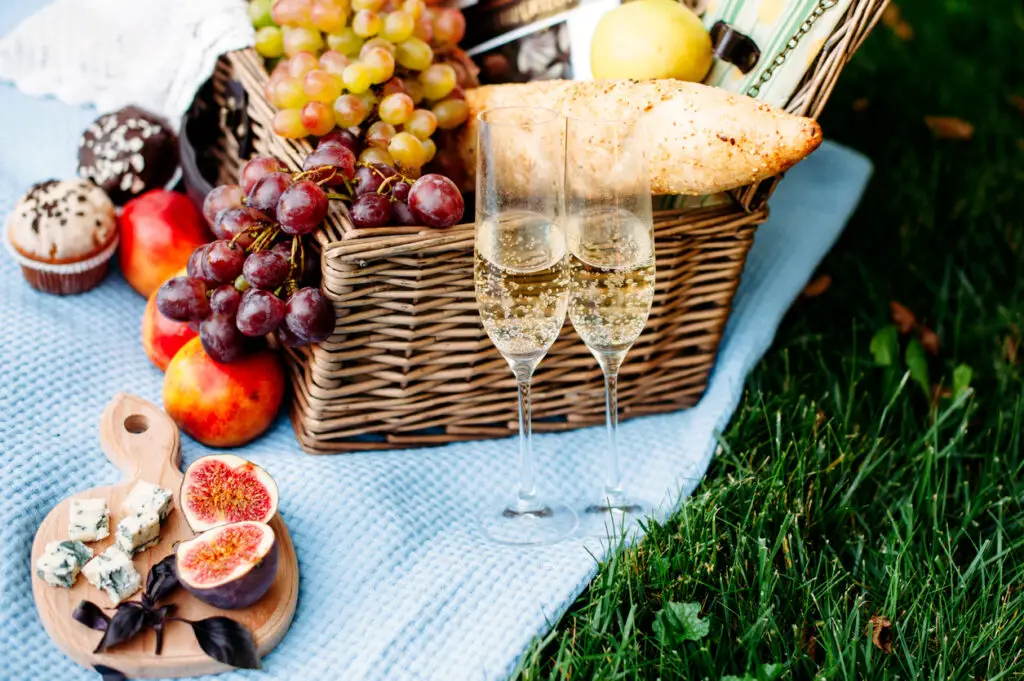 Picnic at the park on a grass, delicious food: basket, wine, grapes, peaches, baguette, cupcakes, figs, cheese, tblue tablecloth, glasses with champagne