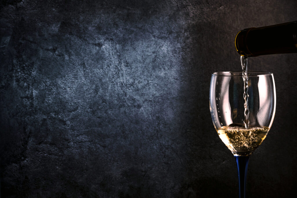 A glass with white wine pouring from a bottle on a dark gray background.
