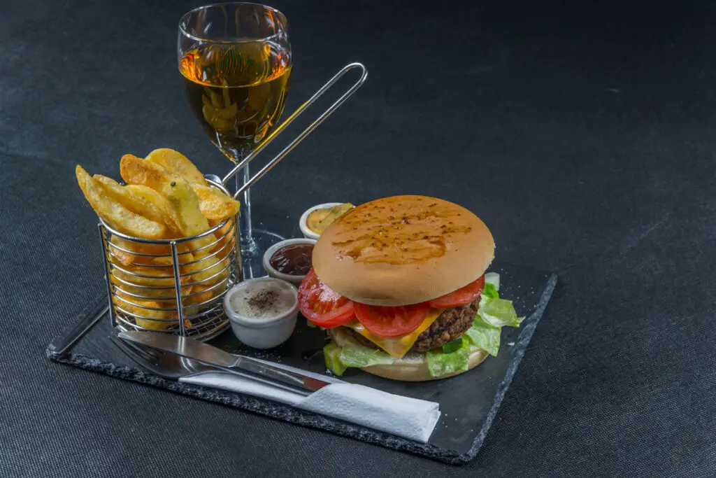 right side view on a flame grilled double stack cheeseburger, lettuce, tomato, with three sauces, chips in a metal basket, white wine