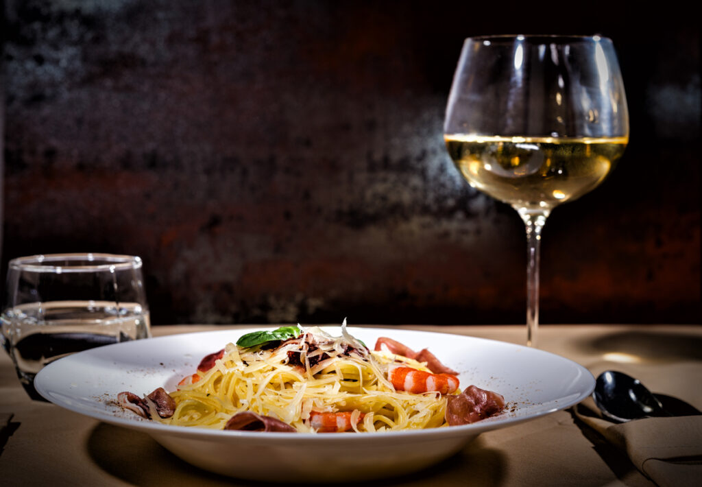 Glass with wine near appetizing spaghetti pasta with shrimps, grated cheese, jamon, tomato sauce and basil in white plate