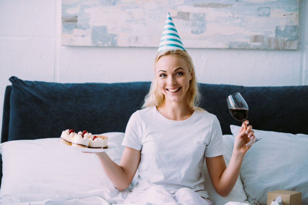 happy woman in party hat holding glass of red wine and cake while celebrating birthday in bed alone
