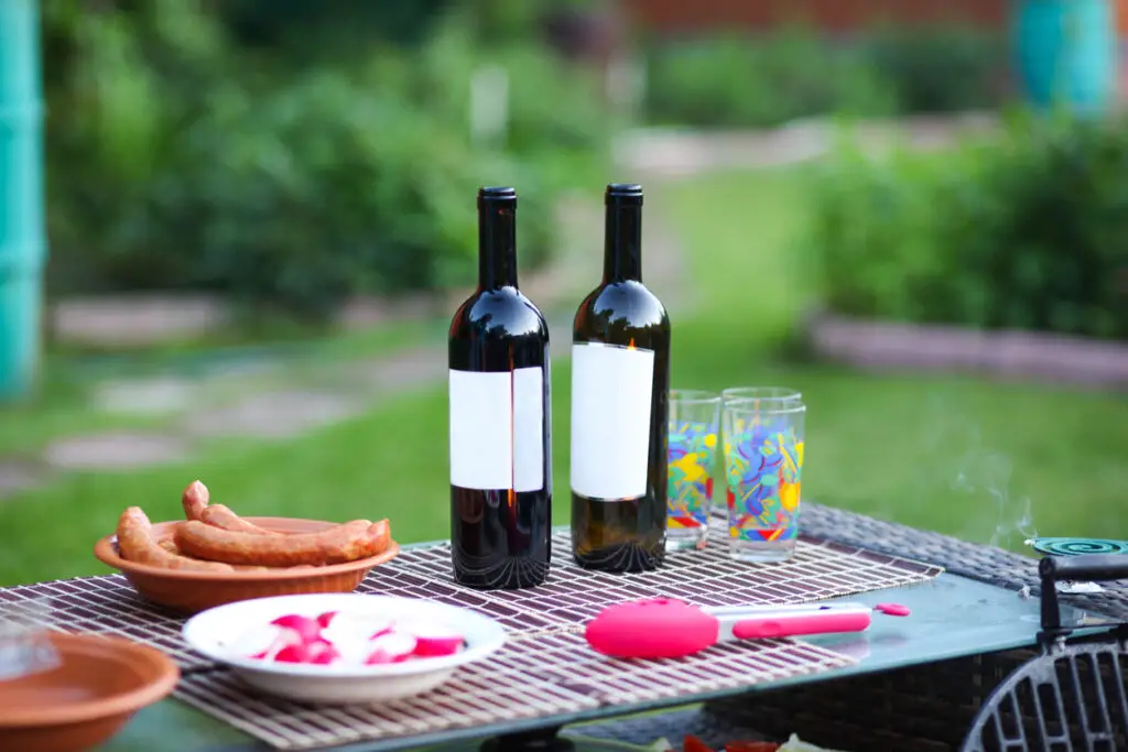 delicious sausages from the grill lie in a plate on a wicker table against the background of green grass. Near two bottles of wine and glasses.