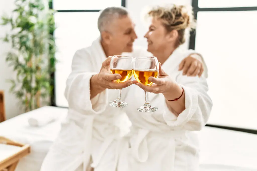Senior caucasian couple smiling happy toasting with champagne sitting on massage table at beauty center.