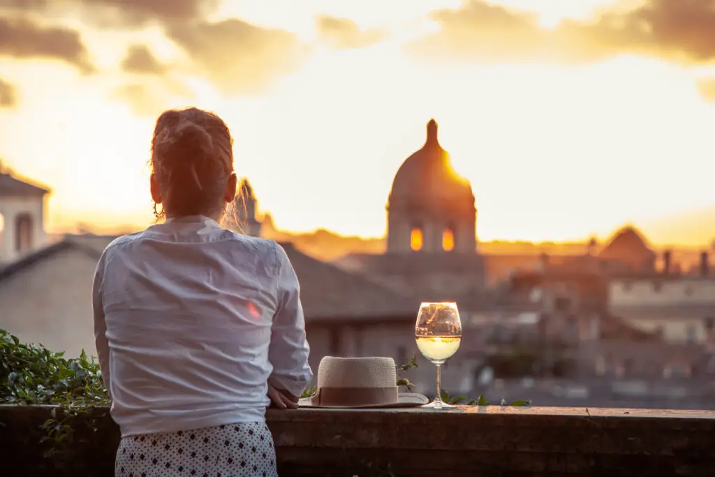 Young woman tourist fashion white dress with glass of white wine in front of panoramic view of Rome cityscape from campidoglio terrace at sunset. Landmarks and domes.