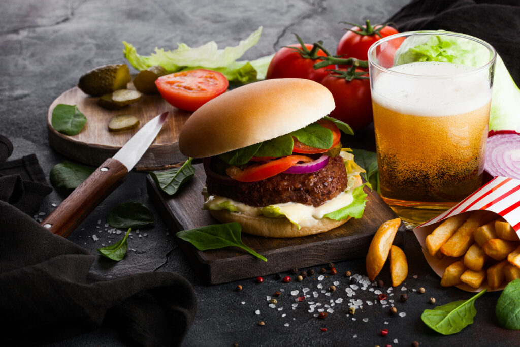 Fresh beef burger with sauce and vegetables and glass of craft lager beer with potato chips fries on stone kitchen table background. Raw organic vegetables on chopping board with knife