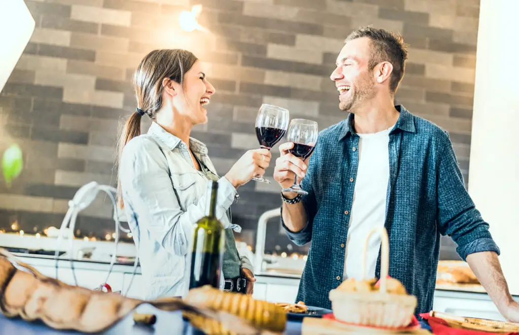 Young couple in love drinking red wine at house kitchen - Happy millenial people at home enjoying aperitif time cheering together at jubilee anniversary - Genuine youth concept on bright indoor filter