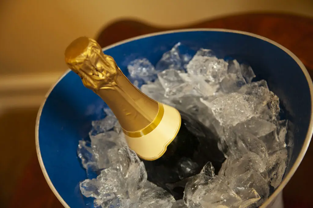 bottle of unopened champagne with gold foil in blue bucket of ice being chilled