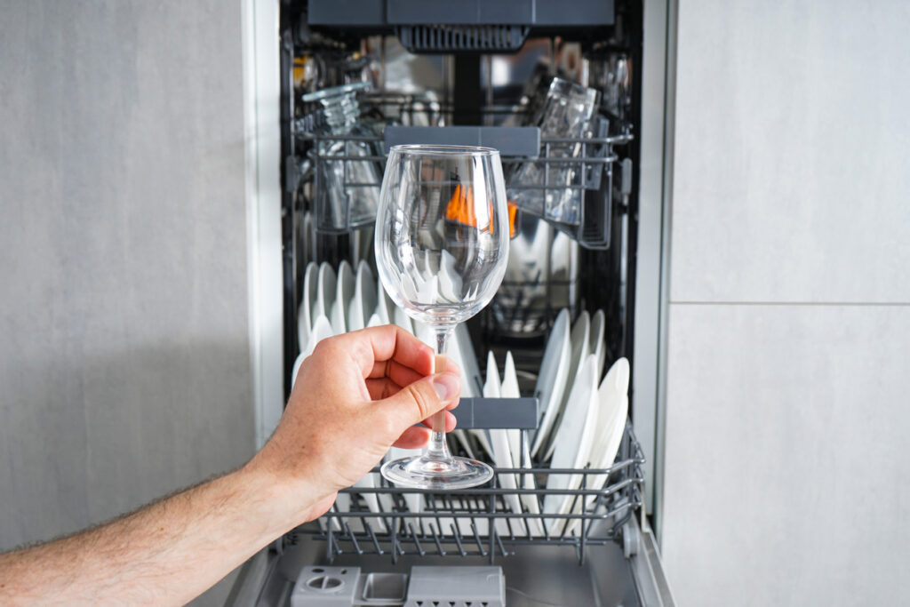 Dishwasher, open and loaded with dishes, front view. Man hand taking out clean wine glass, after washing.